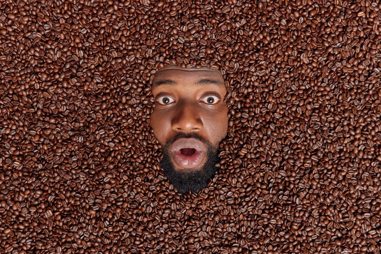 Shocked bearded dark skinned Afro American man buried in roasted coffee beans keeps mouth opened from amazement going to make tasty aromatic beverage from brown seeds. Overhead creative shot